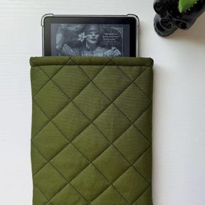 May include: Olive green quilted e-reader sleeve. The sleeve has a diamond pattern with black stitching. An e-reader is partially visible inside the sleeve, displaying a book cover. A black skull-shaped planter with a small succulent is in the background.