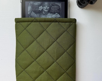 May include: Olive green quilted e-reader sleeve. The sleeve has a diamond pattern with black stitching. An e-reader is partially visible inside the sleeve, displaying a book cover. A black skull-shaped planter with a small succulent is in the background.