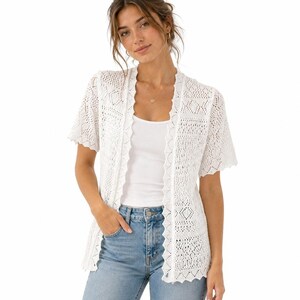 May include: A white short-sleeved open-front cardigan featuring an intricate crochet knit pattern with a scalloped edge along the front opening and hem, worn over a plain white tank top and light blue denim jeans with a visible metal button.