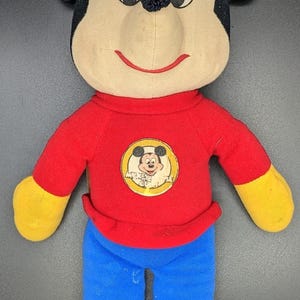 A plush toy of Mickey Mouse wearing a red shirt and blue pants.