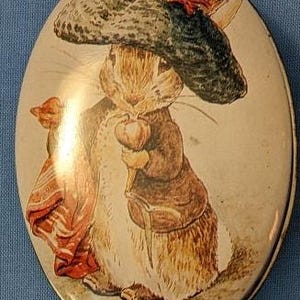 Puede incluir: Una lata ovalada vintage con un conejo de dibujos animados que lleva un sombrero azul y verde con una pluma roja. El conejo sostiene una zanahoria y está de pie sobre un fondo marrón.