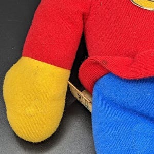 A close-up view of a plush toy, likely a character from a children's television show or movie, wearing a red shirt and blue pants.