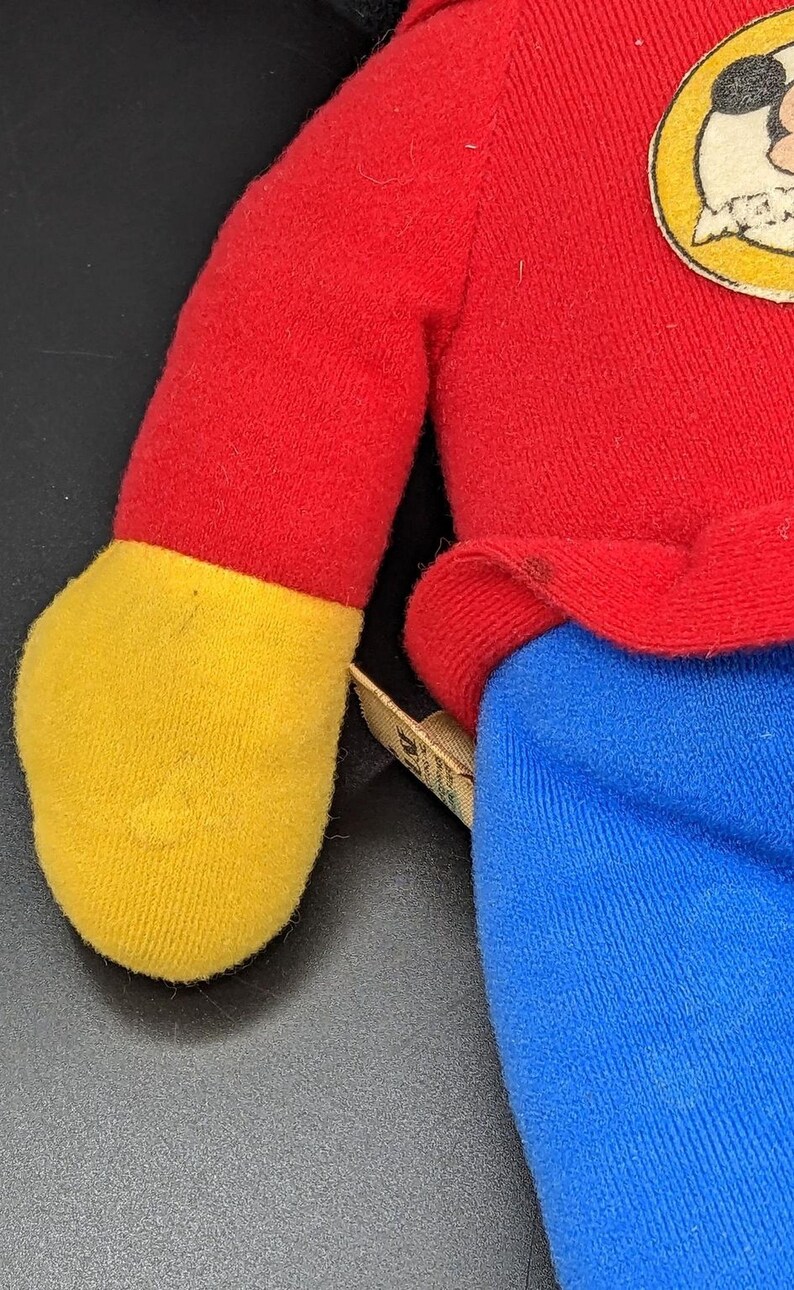 A close-up view of a plush toy, likely a character from a children's television show or movie, wearing a red shirt and blue pants.
