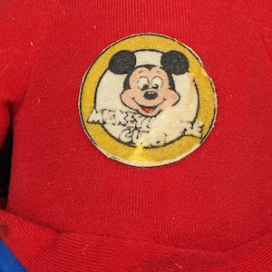 A red sweater with a circular patch featuring a cartoon character, likely Mickey Mouse, on it.