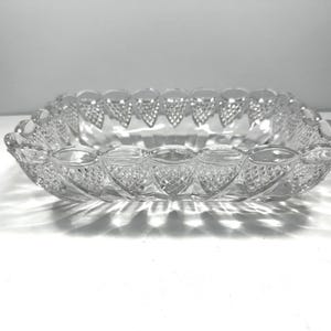 May include: A rectangular clear glass dish featuring a decorative scalloped rim and a textured diamond-cut pattern along the exterior sides. The glass is transparent and reflects light, creating a series of shadows beneath the base.