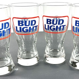 May include: Four clear Bud Light beer glasses. Each glass has a white and red rectangular label with the words "Bud Light" in blue. The glasses are tall and have a curved shape.