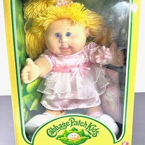 May include: A Cabbage Patch Kids doll in its original packaging. The doll features yellow yarn hair, blue eyes, and a pink dress with a white skirt. The box is yellow and green, displaying the Cabbage Patch Kids logo and the text "I'm One of a Kind, I'm Adoptable!"