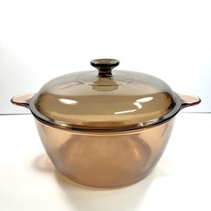 Op de afbeelding: Een ronde ovenschaal van amberkleurig glas met een bijpassend glazen deksel. De schaal is voorzien van twee geïntegreerde handgrepen aan de zijkanten en een centrale knop op het deksel. Het glas is doorschijnend en laat licht door het bruine materiaal.