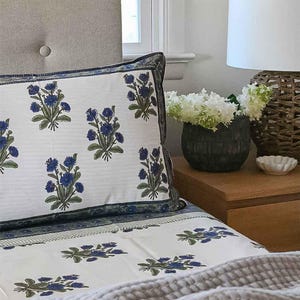 May include: A white pillowcase and sheet set with a blue floral pattern. The bedding features a repeating design of blue flowers and green leaves. The pillowcase has a dark blue border. A gray blanket is at the foot of the bed.