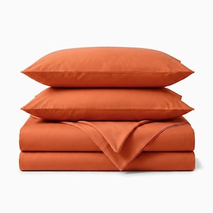 May include: A stack of orange bedding, including two pillowcases and a folded sheet, sits atop a flat sheet. The bedding is a solid, warm orange color, and the items are neatly arranged against a white background.