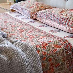 May include: Bedding set featuring a floral pattern in red, green, and gray on a cream background. The set includes two pillowcases and a fitted sheet. A gray quilted blanket is draped over the bed.