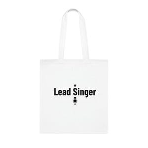 May include: A white tote bag featuring the text "Lead Singer" in black, accompanied by a star and a microphone icon. The bag has two white handles and is made of a durable material. The design is centrally placed on the front.