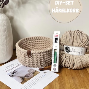 Könnte beinhalten: Ein beigefarbener Häkelkorb mit braunem Lederriemen, eine Kugel beigefarbener Wolle, eine Häkelnadel und Anleitungen. Der Text "DIY-SET HÄKELKORB" befindet sich in einem Kreis im Hintergrund. Auf einer Holzoberfläche.
