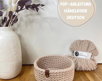 Puede incluir: Una cesta de ganchillo beige con una etiqueta de cuero marrón se muestra junto a una madeja de hilo beige. La cesta redonda tiene un aspecto texturizado. La imagen también incluye un jarrón blanco con flores secas y el texto "PDF-ANLEITUNG HÄKELKORB DEUTSCH."