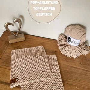 May include: Two beige knitted washcloths with brown leather tags and a ball of beige yarn. The image also includes a decorative metal heart and the text "PDF-ANLEITUNG TOPFLAPPEN DEUTSCH".