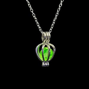 May include: A silver-colored pendant necklace with a cage-like design holding a bright green orb. The chain is delicate, and the pendant has a detailed, openwork structure. The background is solid black, highlighting the necklace.