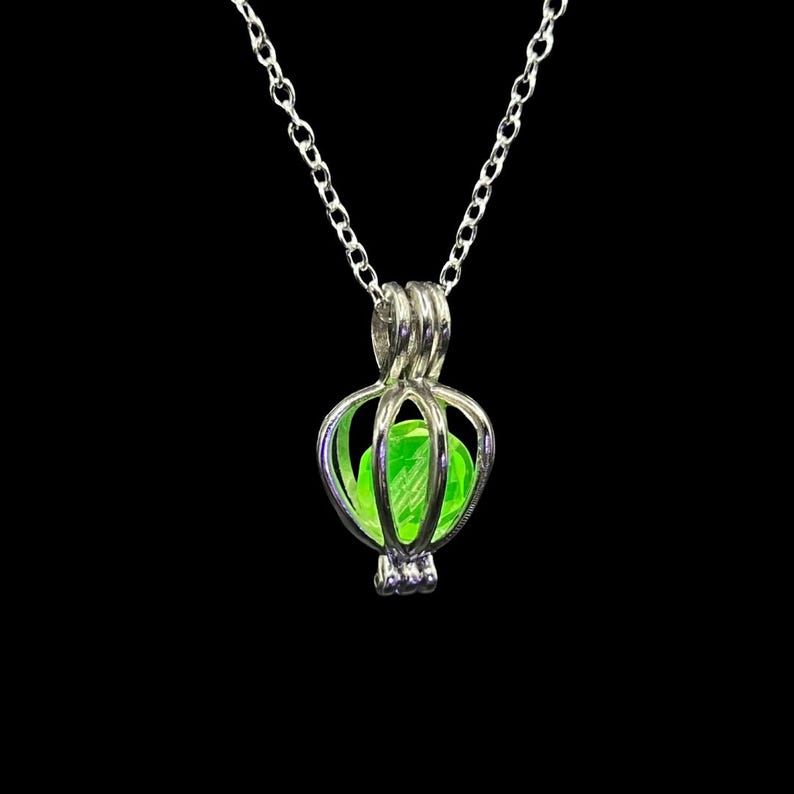 Puede incluir: Un collar con colgante plateado con un dise&ntilde;o en forma de jaula que contiene un orbe verde brillante. La cadena es delicada y el colgante tiene una estructura calada detallada. El fondo es negro liso, resaltando el collar.
