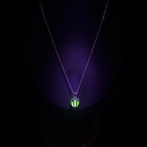 Puede incluir: Un collar plateado con un colgante verde brillante. El colgante tiene forma esf&eacute;rica con un dise&ntilde;o de jaula, y la cadena es delicada. El fondo es negro y el colgante brilla intensamente.