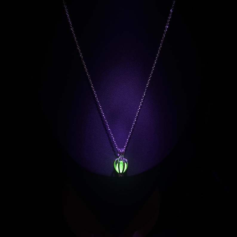 Puede incluir: Un collar plateado con un colgante verde brillante. El colgante tiene forma esf&eacute;rica con un dise&ntilde;o de jaula, y la cadena es delicada. El fondo es negro y el colgante brilla intensamente.