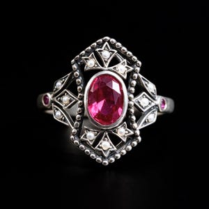 May include: A silver ring featuring a central oval-cut vibrant pink gemstone surrounded by a decorative frame of small white pearls and intricate metalwork. The band includes two small pink stones on the sides, set within a detailed silver structure.
