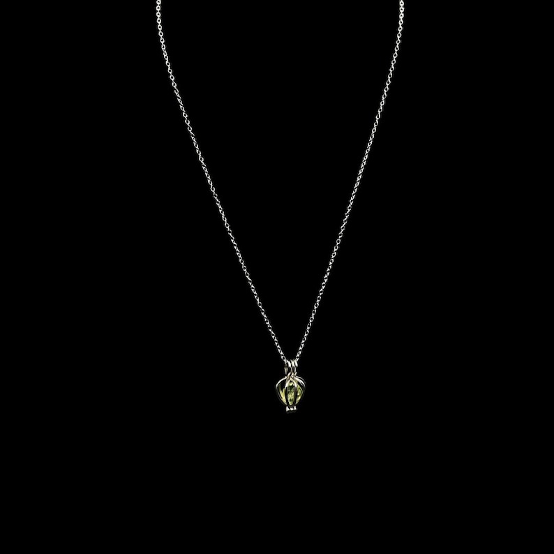 Puede incluir: Un delicado collar de plata con un peque&ntilde;o colgante en forma de jaula que contiene una piedra preciosa verde claro. El colgante cuelga de una fina cadena sobre un fondo negro, resaltando el intrincado dise&ntilde;o y el contraste de color de la joya.