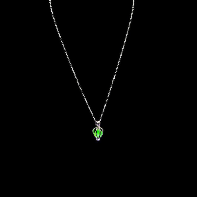Puede incluir: Un collar plateado con un peque&ntilde;o colgante en forma de jaula que contiene una piedra verde brillante. El colgante cuelga de una fina cadena sobre un fondo negro. El collar es una joya sencilla y elegante.