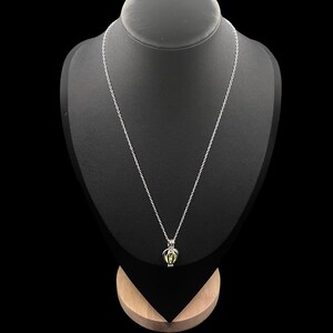 Puede incluir: Un collar plateado con un colgante. El colgante tiene un dise&ntilde;o de jaula, que contiene una piedra preciosa verde claro. El collar se muestra en un maniqu&iacute; negro sobre un fondo negro. La cadena es fina y cuelga.