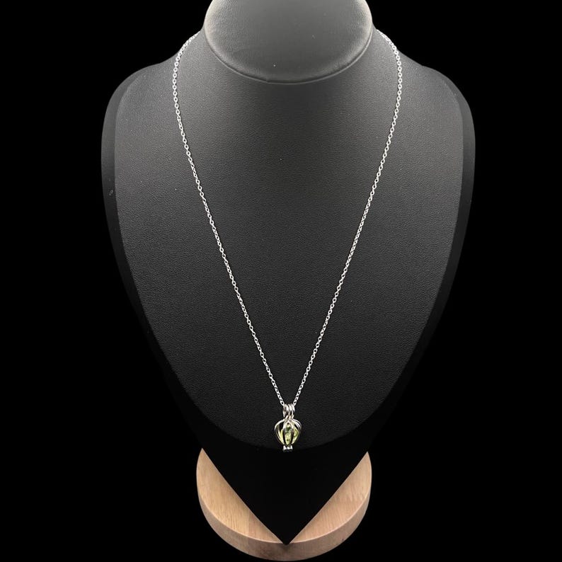 Puede incluir: Un collar plateado con un colgante. El colgante tiene un dise&ntilde;o de jaula, que contiene una piedra preciosa verde claro. El collar se muestra en un maniqu&iacute; negro sobre un fondo negro. La cadena es fina y cuelga.