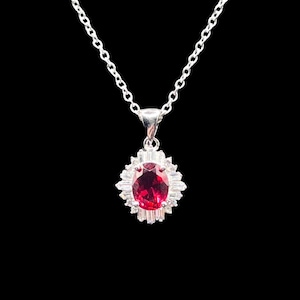 May include: A silver necklace featuring an oval-cut deep red gemstone pendant surrounded by a halo of clear baguette-cut stones. The pendant is suspended from a delicate silver chain with small circular links, creating a classic jewelry piece.