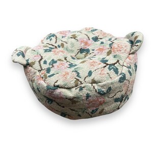 May include: A round, floral patterned cushion with a button in the center. The cushion has a light beige background with pink and green floral designs.