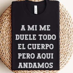 May include: Black t-shirt with white text that reads "A MI ME DUELE TODO EL CUERPO PERO AQUI ANDAMOS." The shirt is displayed on a round, woven, beige surface. The shirt has a crew neck and a tag.