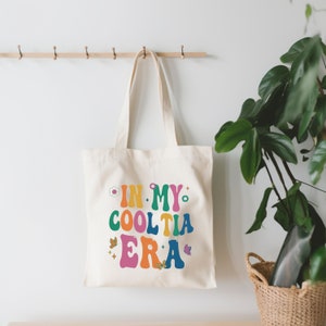 Op de afbeelding: Cr&egrave;mekleurige draagtas met de tekst "IN MY COOL TIA ERA" in kleurrijke letters in retrostijl. De tas heeft een bloemen- en vlinderontwerp. De tas hangt aan een houten rek.