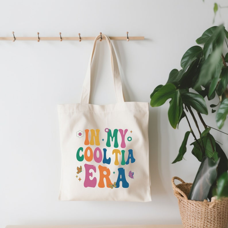 Op de afbeelding: Cr&egrave;mekleurige draagtas met de tekst "IN MY COOL TIA ERA" in kleurrijke letters in retrostijl. De tas heeft een bloemen- en vlinderontwerp. De tas hangt aan een houten rek.