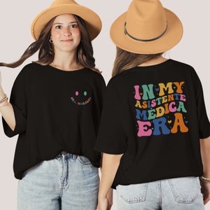 May include: Black t-shirt with a smiley face on the front and the text "IN MY ASISTENTE MÉDICA ERA" in rainbow colors on the back. The text is surrounded by small hearts.
