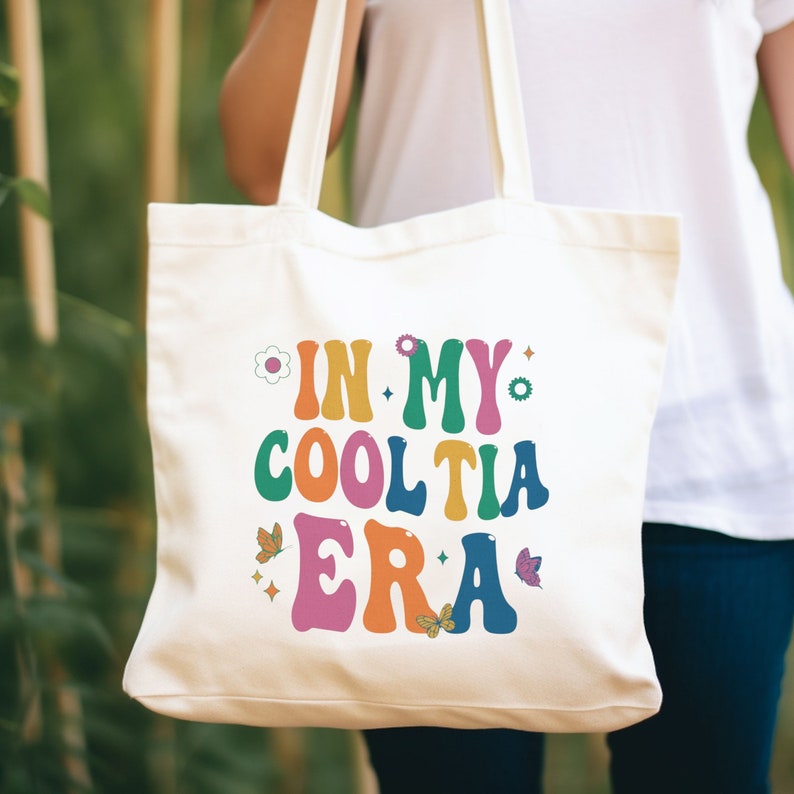 Op de afbeelding: Cr&egrave;mekleurige draagtas met de tekst "IN MY COOL TIA ERA" in kleurrijke, retro-stijl letters. Het ontwerp bevat bloemen- en vlindergraphics. De tas wordt door een persoon vastgehouden.