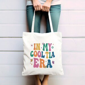 Op de afbeelding: Een witte draagtas met de tekst "IN MY COOL TIA ERA" in kleurrijke, retro-stijl letters. Het ontwerp bevat bloemen- en vlindergraphics. De tas wordt vastgehouden door een persoon die een blauwe spijkerbroek draagt.