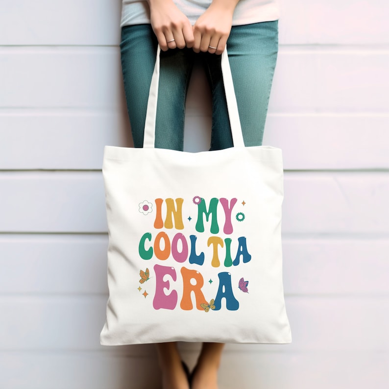 Op de afbeelding: Een witte draagtas met de tekst "IN MY COOL TIA ERA" in kleurrijke, retro-stijl letters. Het ontwerp bevat bloemen- en vlindergraphics. De tas wordt vastgehouden door een persoon die een blauwe spijkerbroek draagt.