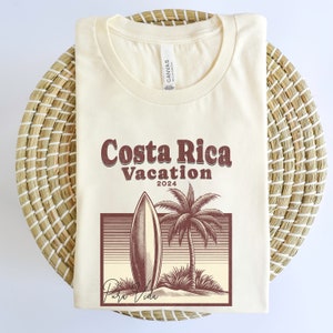 May include: A beige t-shirt with a vintage-style graphic design featuring a surfboard, palm tree, and the text "Costa Rica Vacation 2024" in brown. The text "Pura Vida" is written in cursive below the graphic.