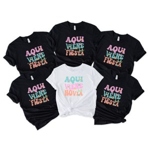 May include: Five black t-shirts with the text "Aqui Viene La Fiesta" in pink, blue, and orange letters. One white t-shirt with the text "Aqui Viene La Novia" in pink, blue, and orange letters.