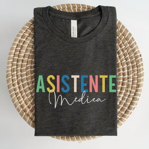 May include: A dark gray t-shirt with the text "ASISTENTE Medica" in colorful letters. The text is in a rainbow color scheme.