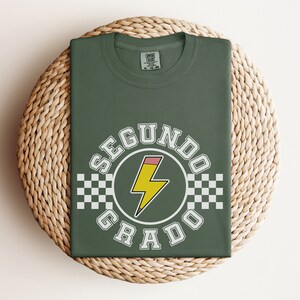 May include: A green t-shirt with a white circle graphic that says "SEGUNDO GRADO" with a yellow lightning bolt inside the circle. The lightning bolt is drawn to look like a pencil with a pink eraser.