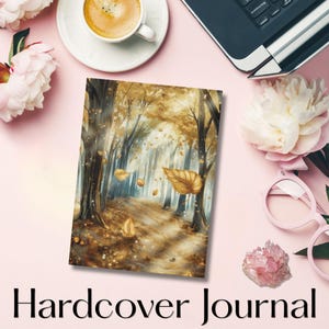 Puede incluir: Diario de tapa dura con una escena de bosque en tonos dorados y marrones, con hojas cayendo. Las palabras "Hardcover Journal" están impresas en la parte inferior. El diario está sobre una superficie rosa con una taza de café, flores y gafas.