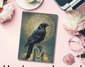 Op de afbeelding: Hardcover journal met een gedetailleerde illustratie van een zwarte raaf die op een kristal zit, tegen een achtergrond van een gouden mandala. Het journal is omgeven door een kop koffie, bloemen en een laptop.