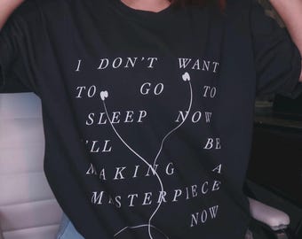 May include: A black short-sleeved t-shirt featuring white text that reads I DON'T WANT TO GO TO SLEEP NOW I'LL BE MAKING A MASTERPIECE NOW, accompanied by a white graphic illustration of wired earbuds with the cord forming a decorative line.