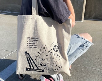 May include: A beige canvas tote bag featuring black line art and the text "STAY ESSENTIALS" with a list of items. The design includes a compass rose, stars, and a heart. The words "SKZ CODE" are also visible.