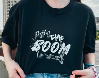 May include: Black t-shirt with the words "CHK CHK BOOM" in a white, comic book style font. The shirt also has the text "VAMOS, I KNOW THAT YOU WANT IT" in white. The shirt is paired with blue jeans.
