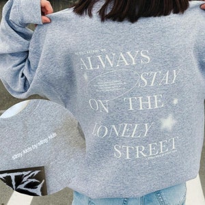 Peut inclure: Un sweat-shirt gris avec du texte blanc au dos : "ALWAYS STAY ON THE LONELY STREET." Le texte est dans une police stylisée avec un motif d'étoile. Les mots "stray kids by stray kids" sont imprimés sur le devant.