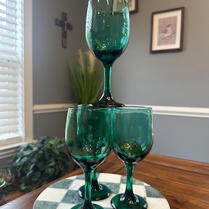 May include: Four emerald green wine glasses are stacked on a marble and checkered serving tray. The glasses have a classic design with a rounded bowl and a long stem. The glasses are arranged on a wooden table.
