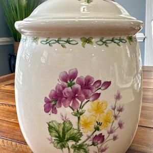 May include: Cream-colored ceramic jar with a lid, adorned with a floral design featuring purple and yellow flowers and green leaves. The jar has a rounded shape and a decorative vine pattern around the rim.