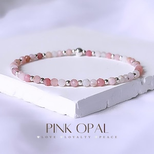 May include: A delicate pink opal bracelet with silver beads. The bracelet is on a white surface and the text "PINK OPAL LOVE LOYALTY PEACE" is below the bracelet.